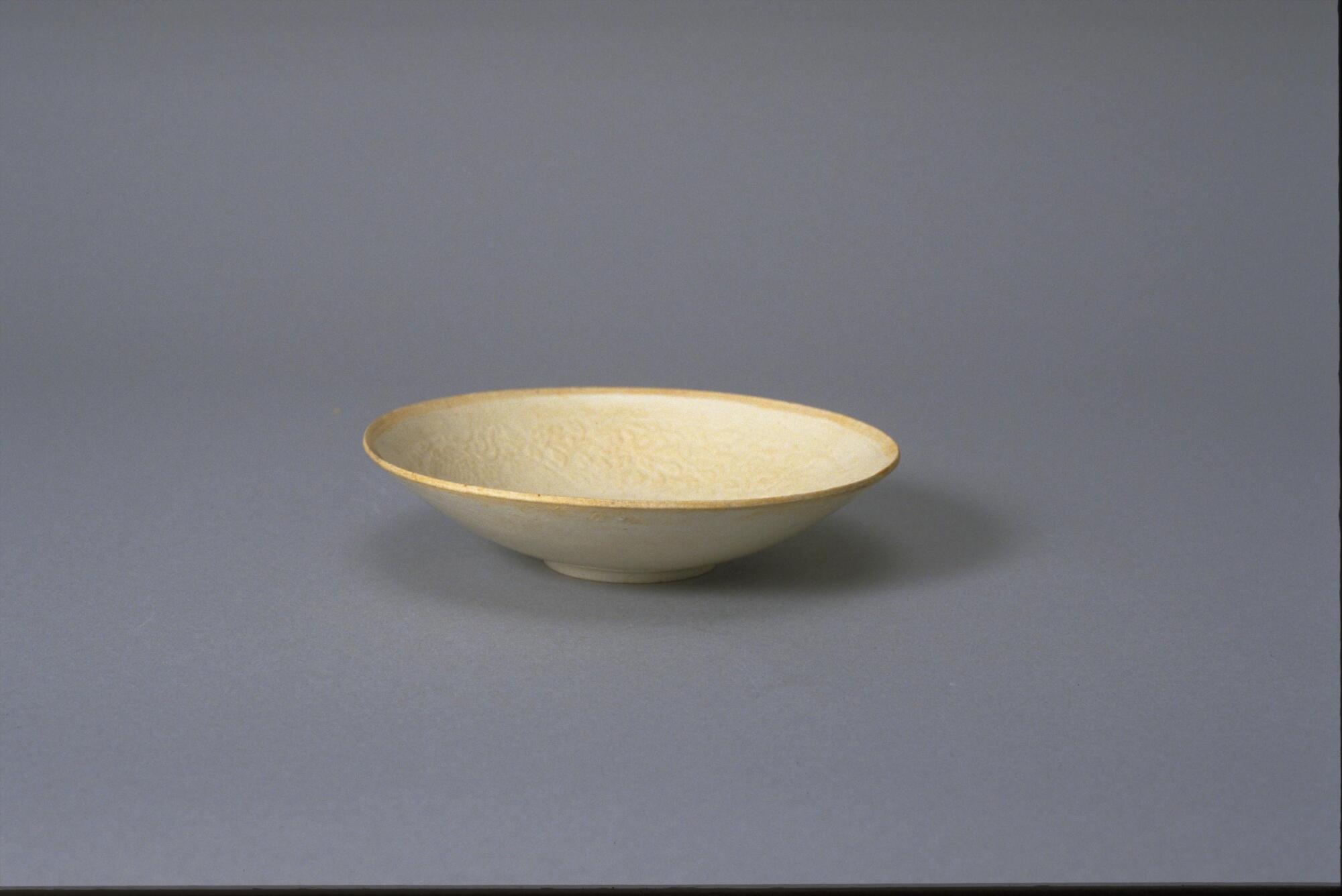 This thin porcelain conical bowl with direct rim on a footring has an interior with incised floral meander decoration. It is covered in a white glaze with bluish tinge and has an unglazed rim.