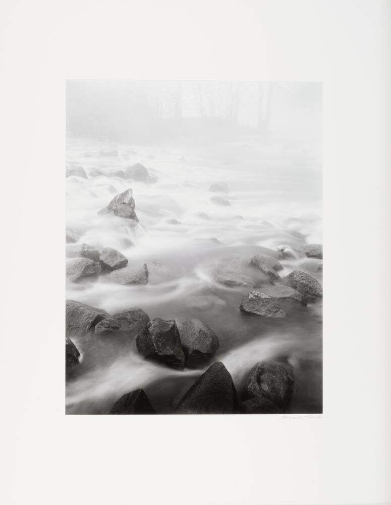 An image of water flowing over large boulders in a river. Photographed with a long exposure, the water appears like bright fog winding its way around a dry bed of rock.&nbsp;