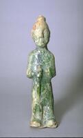 A red earthenware figure of a Chinese man, standing with hair in a top knot, wearing a long robe over pants, and holding a weapon on a long stick in front of him. The figure is covered in a green lead glaze, with iridescence and calcification.