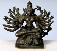 Durga sits with her legs in a half lotus position, crossed in front of her, but not interlaced.  She has a narrow waist and rounded pointy breasts with broad shoulders.  Her front two hands hold a rosary (also in a reassuring gesture) and a pot. Her other hands fan out around her. Reading clockwise, she carries a wide assortment of weapons, an arrow, sword, feather, club, discus, trident and [?] on her right and conch, bell, noose, trident, club?, shield, bow and a kapala (skull cup).  She has large open eyes and a full mouth and nose.  She wears jewelry including necklaces and shoulder loops, armlets, bracelets and large floral earrings.  Her crown rest atop her head, but there are wing-like elements that fan out behind her ears.  She sits on a squared base with stylized lotus petals over simpler moldings. <br />