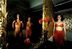 The work is a color photograph of three models in red lingerie. Two models are visible only in the reflection of a large, ornately-framed wall mirror. A third model stands to the right, surrounded by floral wallpaper. A vase of red calla lillies sits on a table in front of the mirror. A single lily is placed on the table and can be seen in the mirror's reflection.