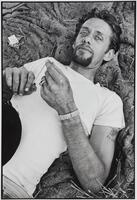 A photograph of a man lying on a blanket in the grass, a pack of cigarettes beside him. He wears a white t-shirt and a band around his wrist. He pulls a string taught between his fingers, while looking into the camera lens. 