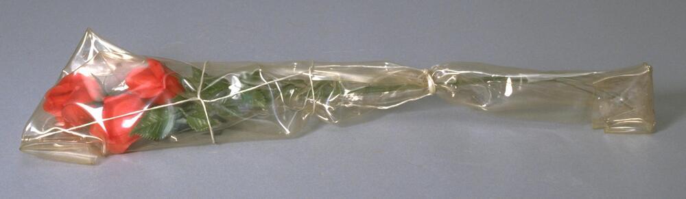 Three plastic long-stemmed red roses wrapped in thick transparent polyethylene, tied with twine, ends stapled