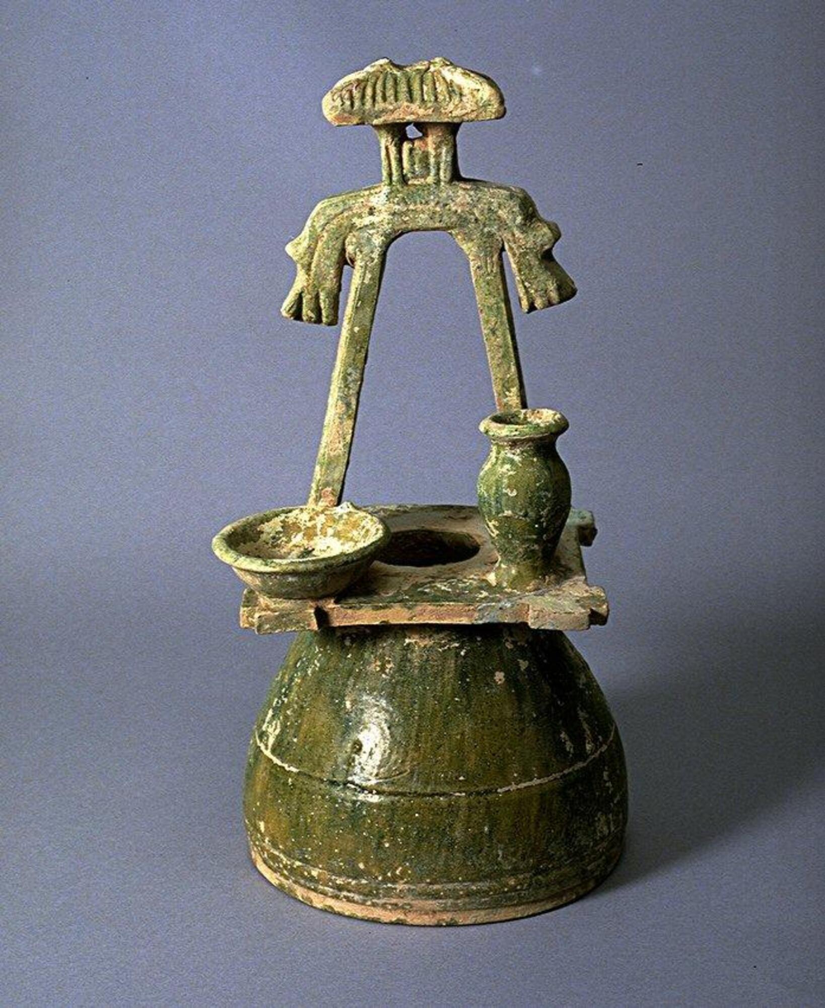 This red earthenware model of a well-head is constructed from an upside-down, wheel-thrown bowl with a hole pierced in its base to create the opening for the well. Its wide rim creates the well-head's base, and a flat clay slab forms the framed opening and windlass. It is topped with a small hip and gabled roof pavilion, and a basin and amphora rest on the framing. The model is covered in a green lead glaze, with iridescence and calcification. 