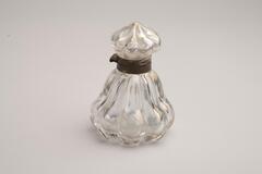 This is a clear glass inkwell with a metal collar. The bottle is pear-shaped with vertical segments that are bulbous at the base. The hollow glass cap has this bulbous pattern that rises to a pointed top. The glass has some iridesence.<br />