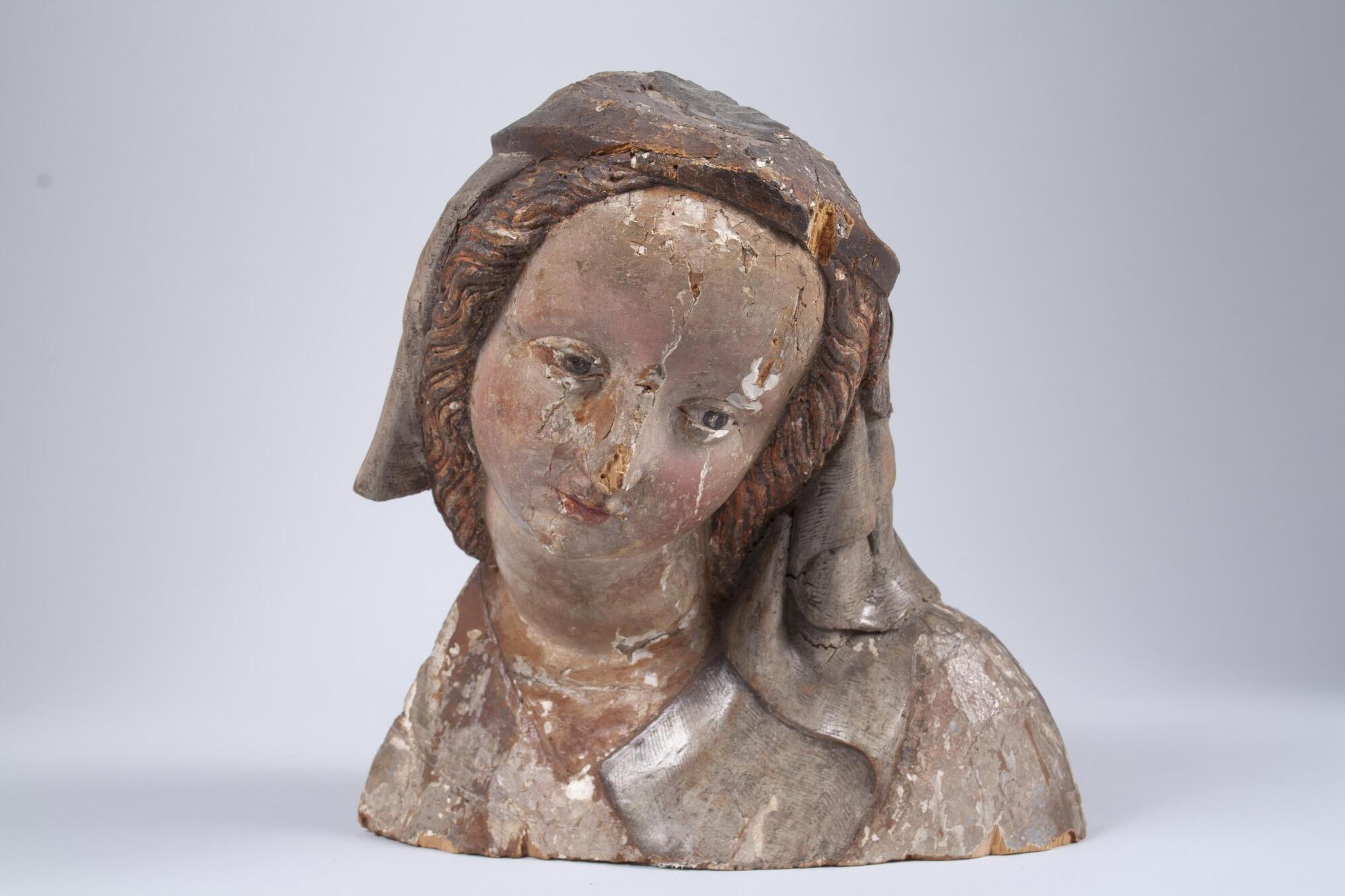 Bust of Madonna with head facing down and to the side. The main sculpture is of wood with layers of paint on the face and veil.