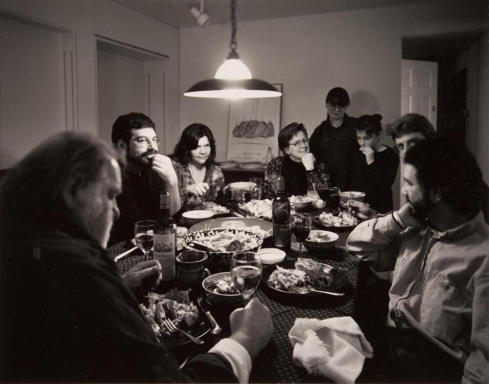 A photograph of a group of people sitting around a dinner table, engaged in conversation. The food is placed on the center of the table. The sitters eat and drink wine.