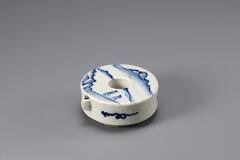 It is simple in shape, like a donut but with a sharply trimmed rim in the manner of a metal vessel. The hole in the middle is believed to be a symbol of Eastern philosophy. Designs are painted on the surface in cobalt blue pigment.<br />
<br />
This ring-shaped water dropper is decorated with a figures-in-landscape design on its upper surface and a floral scroll design on its sides rendered in cobalt blue. A line runs around the foot and sand was used as kiln spurs. The clay and glaze are well fused. This is one of many water droppers that were produced at Bunwon-ri, Gwangju-si, Gyeonggi-do, in the late 19th century.<br />
[Korean Collection, University of Michigan Museum of Art (2014) p.183]