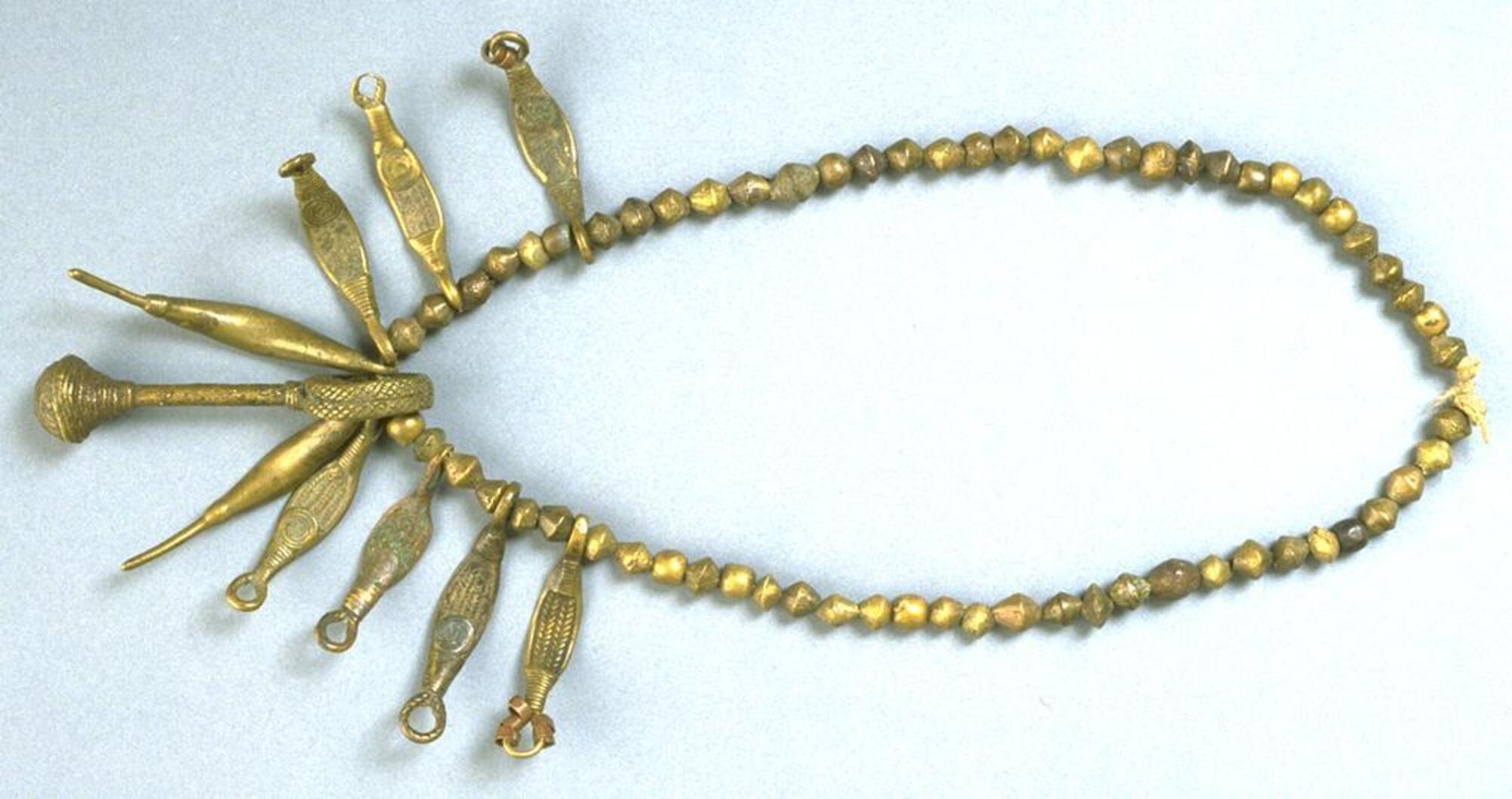 Belt made of brass beads with brass pendants. The central pendant is in the form of a crotal bell attached to a short rod with a large suspension loop. Two pendants are curved forms and the other seven are in the shape of what have been described as banana leaves. 