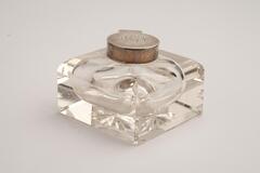 This is a cut crystal glass inkwell in the shape of a square. The upper edges are rounded and there are designs cut into the bottom. The round cap is silver with a monogram design.<br />