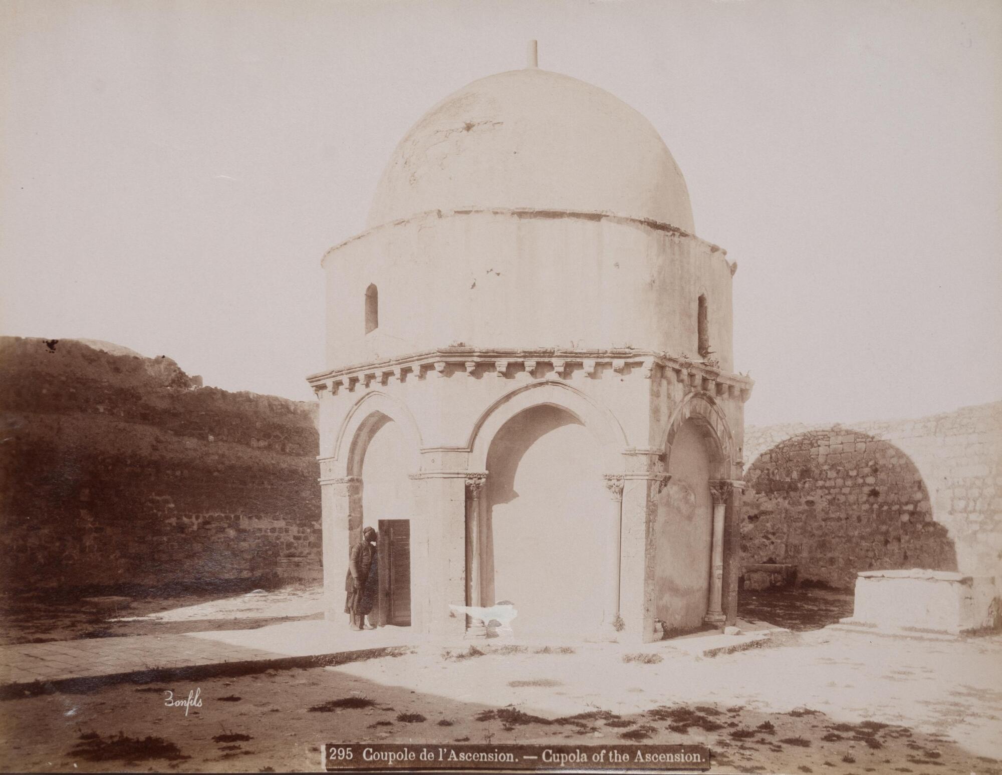 This photograph depicts a prominently placed domed structure in the center of a courtyard with a man standing at the threshhold of the door. 