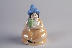 A porcelain inkwell of a woman sitting. She is holding a green vase in both hands and is wearing a blue pointy hat. The rest of her outfit is orange.
