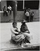 Two women sit in chairs next to a large metal pole at a street curb. Men, women, and children sit on a stone wall same distance from the street.