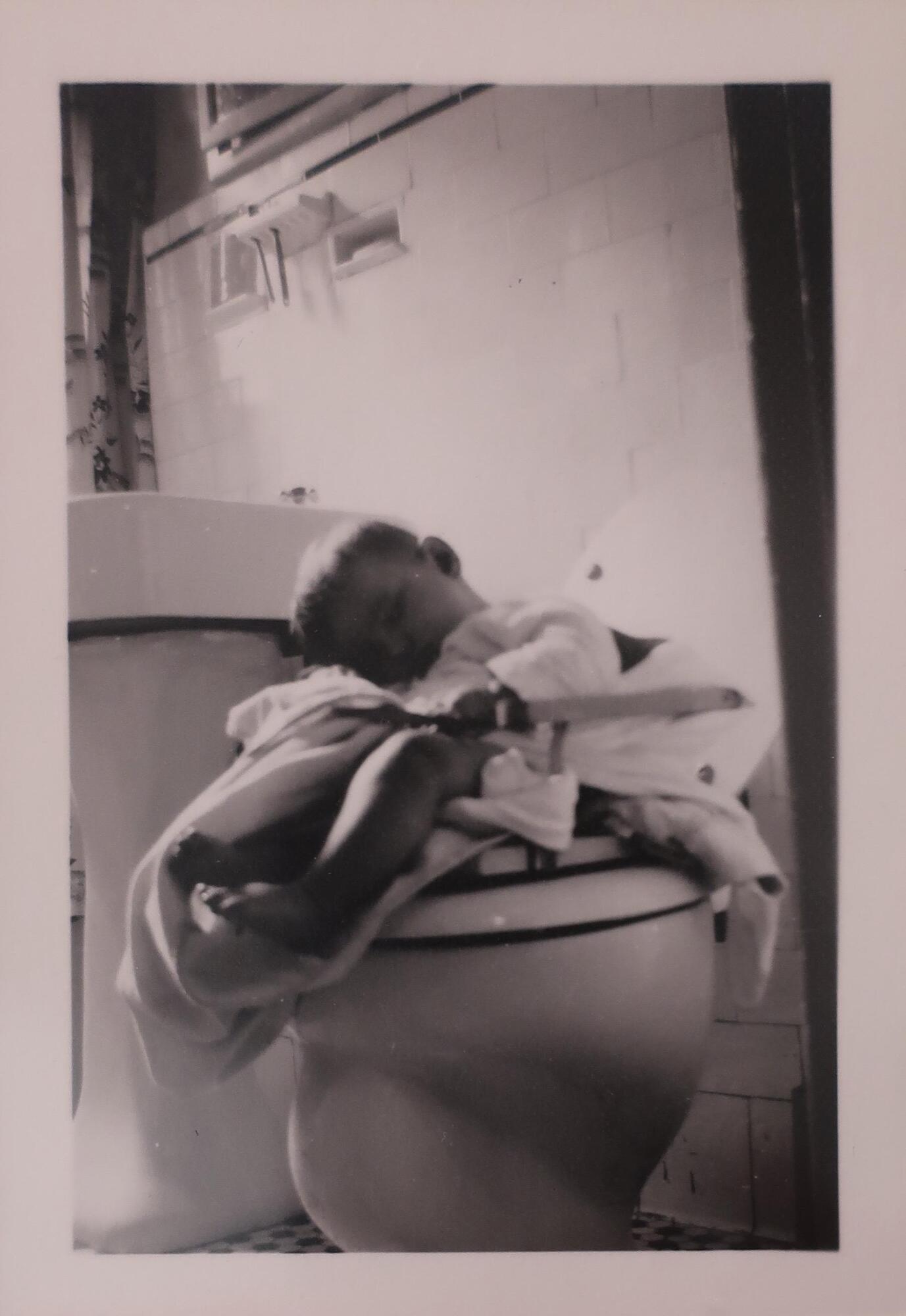 <span style="font-size:12pt"><span calibri="" style="font-family:">A small child sleeping in a chair on what appears to be the lid of a toilet. The child has blankets around them and is leaning over to the left. There is white tile and a wall-mounted toothbrush holder visible in the background.</span></span><br />
&nbsp;