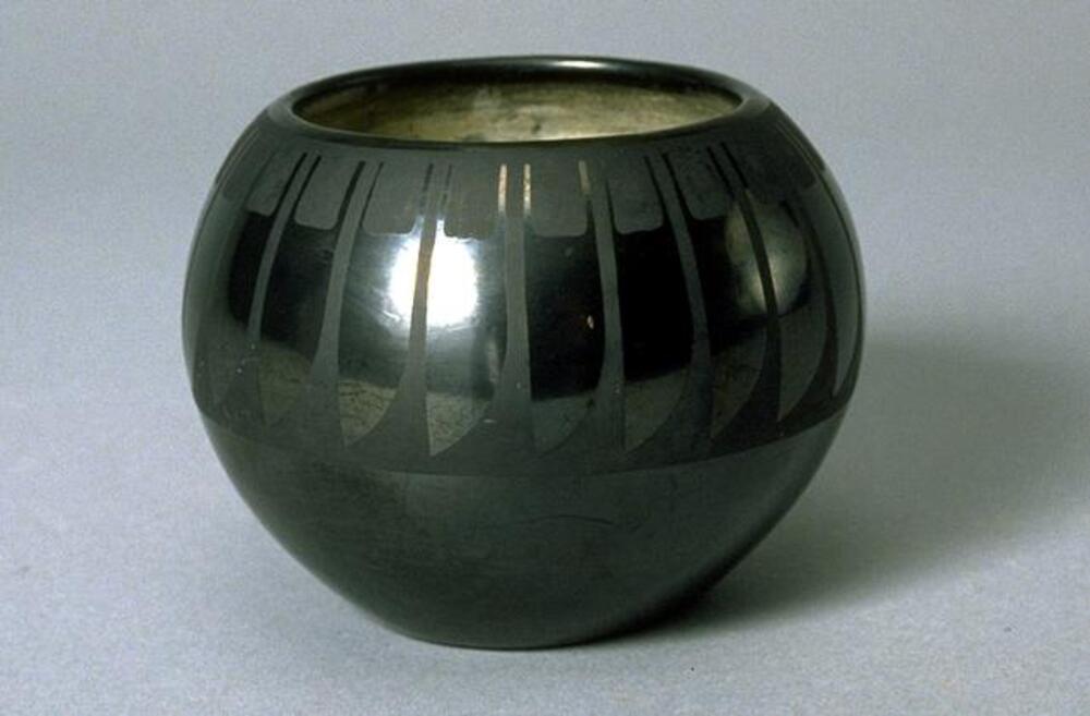 This small spherical pot with wide mouth is decorated in black-on-black style. Around the upper half is a feather design, which looks like individual feathers hanging down from the mouth forming a ring around the circumference. 