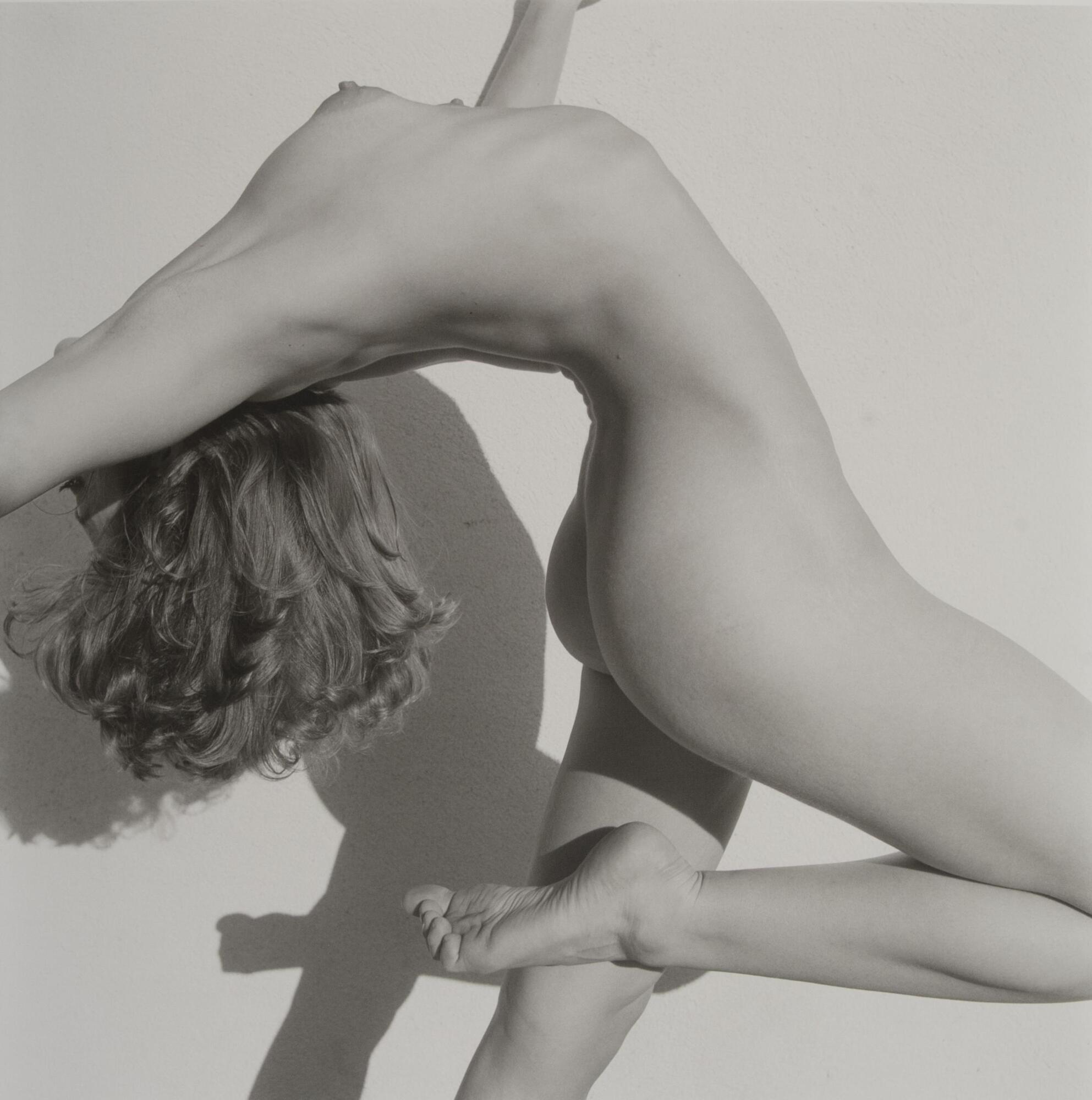 A nude in a dance pose. The female dancer is posed with an arched back and her arms extended, one knee is brought up as if in an exaggerated step motion. Her head is tilted back with her face covered and hair hanging down. A light source to her right casts a shadow of her body onto a white background.