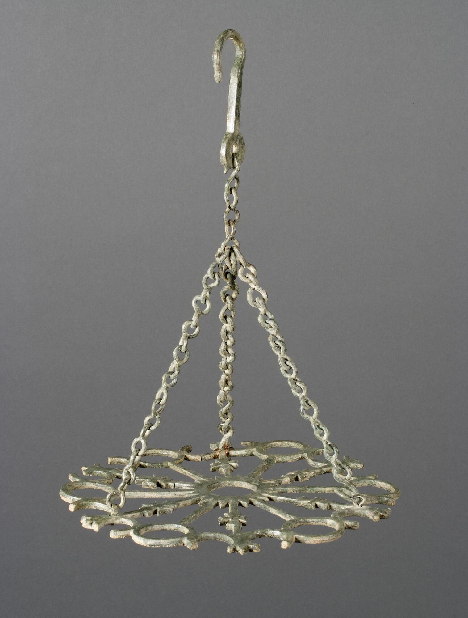 This bronze light fixture consists of a central circular celestial motif from which extend twelve arms in a radial pattern. Six of these arms, embellished with maltese crosses, end in omega-shaped terminals. These decorated arms alternate with six unadorned arms that terminate in rings designed to hold glass oil lamps. The entire disk is suspended from three bronze chains joined to a large hook.