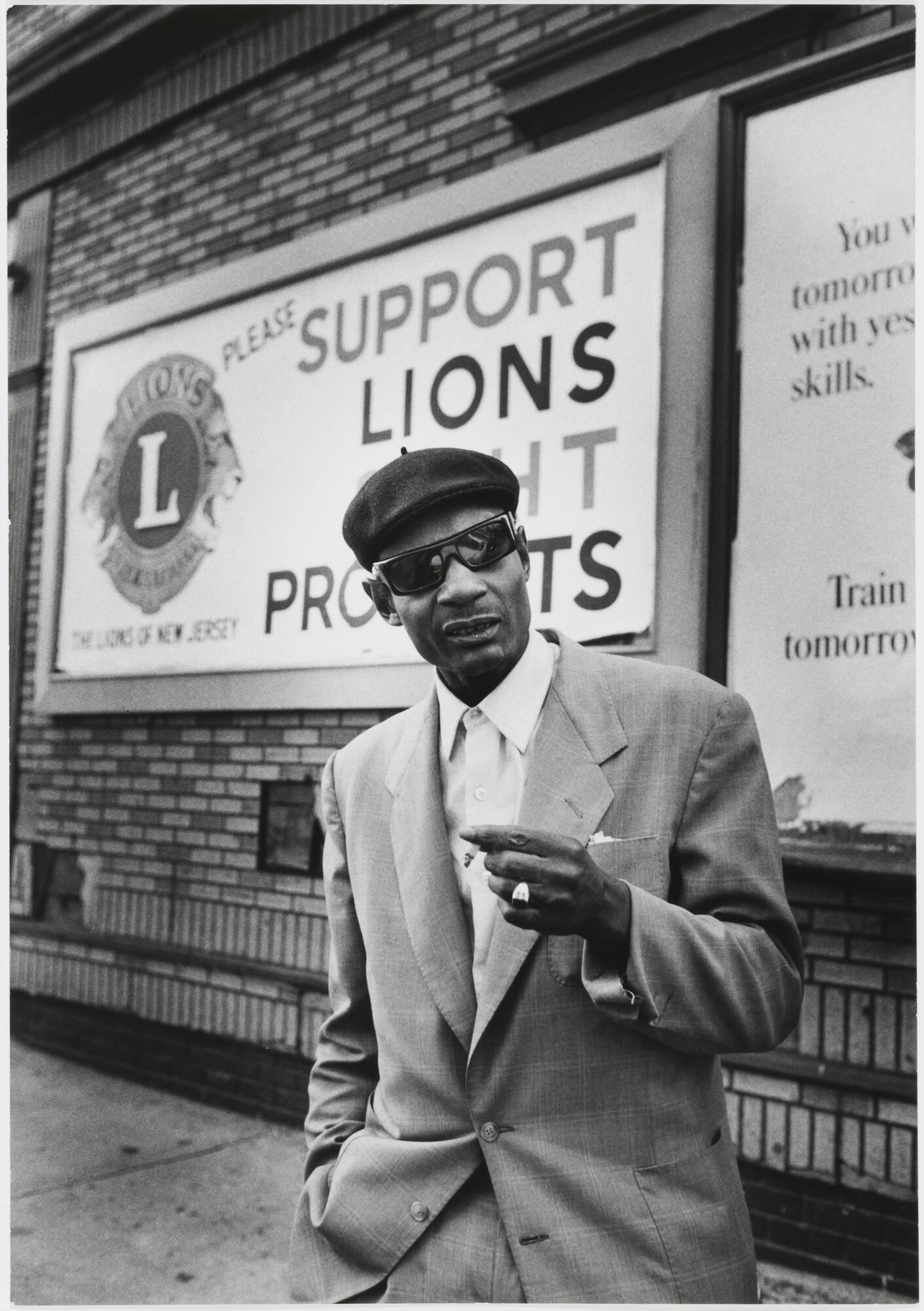 Image of a man standing in front of a brick building with an advertisement for &quot;The Lions of New Jersey.&quot; He wears a light-colored window pane suit,sunglasses, and dark beret. His ringed left hand holds a cigarette.&nbsp;