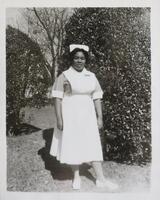 A woman in an old-fashioned nurse&#39;s uniform standing in front of two large bushes. She is smiling slightly, posed with her left foot turned out and her arms at her sides.