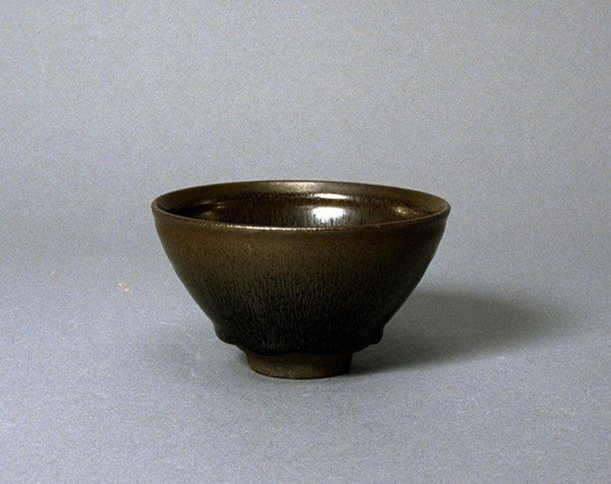 A deep, conical bowl on a straight foot ring and with subtle rim articulation, covered in a thickly applied dark iron-rich black glaze with lighter russet-brown and iridescent hare's fur (兔毫盏 <em>tuhao zhan</em>) markings.  The thick glaze thins at the rim to a russet-brown color.