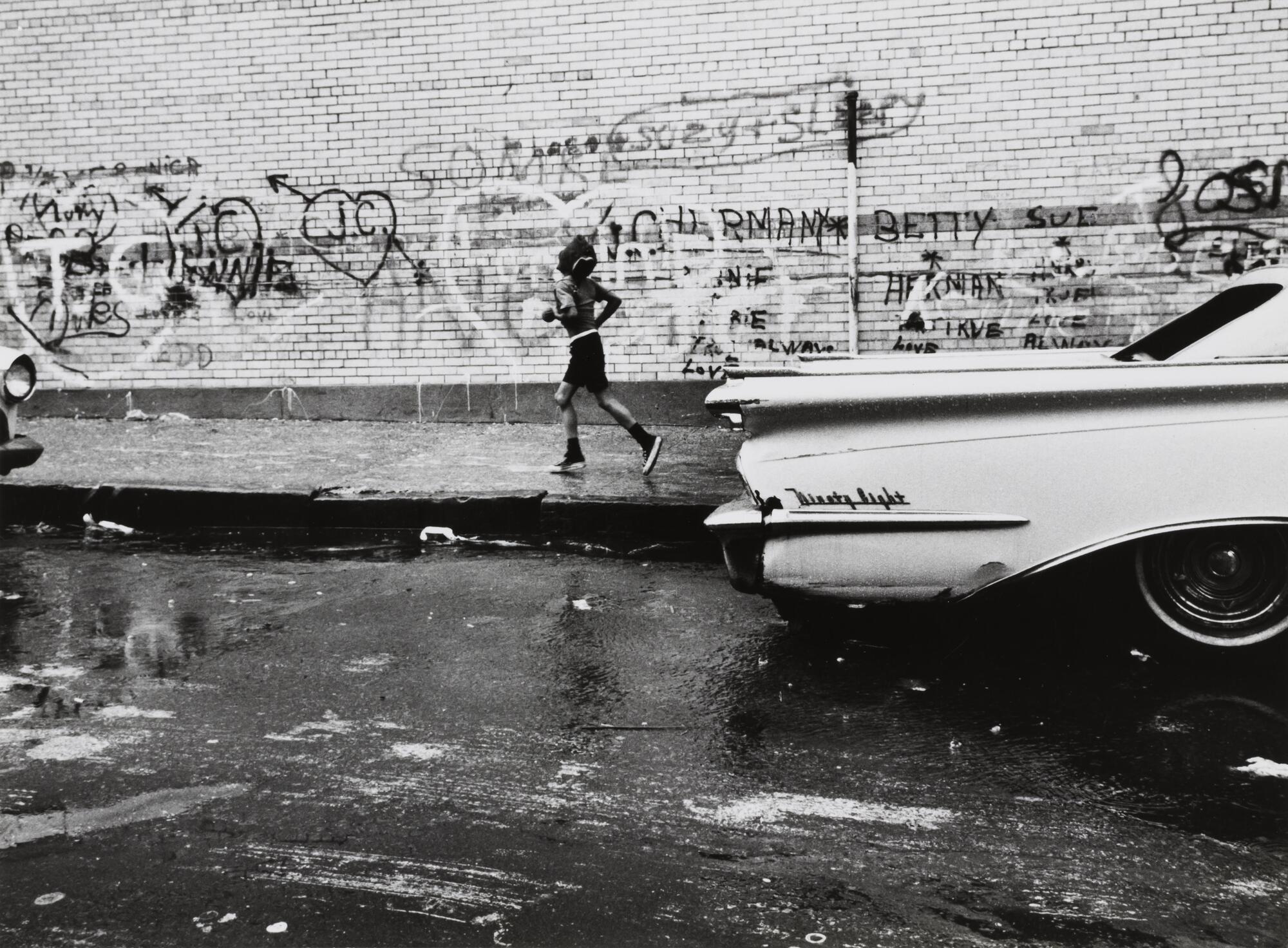 A boy in shorts runs along the side of a graffiti- covered brick building. He passes a parked car with tail fins.