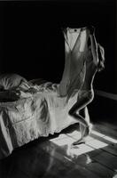 This photograph shows a young girl holding a garment above her head in a domestic space. She stands in a spot of light pouring in from a window, next to an unmade bed.