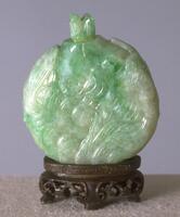 A circular sea foam green and white jadeite snuff bottle with fish and waves incised on the surface. The snuff bottle stopper is shaped like a fish and the snuff bottle is sitting on an incised wooden stand.