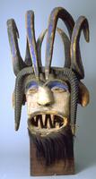 Wooden mask with a superstructure of curling horns with blue pigment. The face of the mask is white and the mouth is open with pointed teeth. Attached below the mouth is a panel of fur, possibly monkey fur. The forehead is dark in color while there is blue pigment above each eye. On each side of the head are possible animal figures or horns, while atop the forehead sits another set of horns or hairstyle that points downward, carved with a spiral design. 
