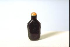 A purple glass snuff bottle with a flower tree branch and inscription carved onto the front. On top of the snuff bottle is a mouthpiece with a green glass collar and an orange carnelian stopper.&nbsp;