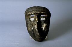 A small, oblong shaped mask with mirrors in the eyes and on the cheeks. The mask is dark in color, with a raised forehead ridge and small slits above the mirror-covered eyes. The nose is long and twists to one side of the face. 