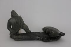 A figure of a man wearing a parka with one leg bent behind him as though he is pulling something heavy. There are small wooden pegs on the bottoms of the man&#39;s feet where the figure attaches to the base of Man Pulling Walrus.&nbsp;
