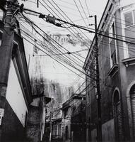 This photograph depicts a view of an urban scene with telephone poles and electrical wires strung up and down a winding street.<br />
 