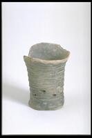 This gray earthenware tall-stemmed pedestal vessel is covered in black burnished slip with a bowstring pattern and a few piercings around the body. There is a flaring rim on a narrow base, and loss of rim.  