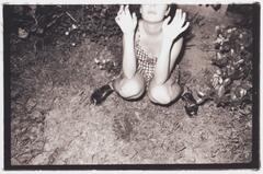 A woman squatting on the ground, hands raised. There are trees, dirt and leaves surrounding her.