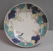 The circular, smaller white porcelain plate has a design of gourds, flowers, vines and leaves around the rim. The gourds are outlined with blue underglaze and colored with yellow overglaze. Blue underglaze and transparent green overglaze are used for the leaves. The flowers and vines are drawn with red. The red enamel is worn off from some of the tendrils, a characteristic of 18th century Nabeshima. The reverse side has four clustered jewel or treasure motifs with four bows and streamers repeated three times. On the shallow foot, bold lines are drawn in a row like a comb. The design on the back is all drawn with blue underglaze. (Referencce: Becker, Sister Johanna. “A Group of Nabeshima Porcelain.")