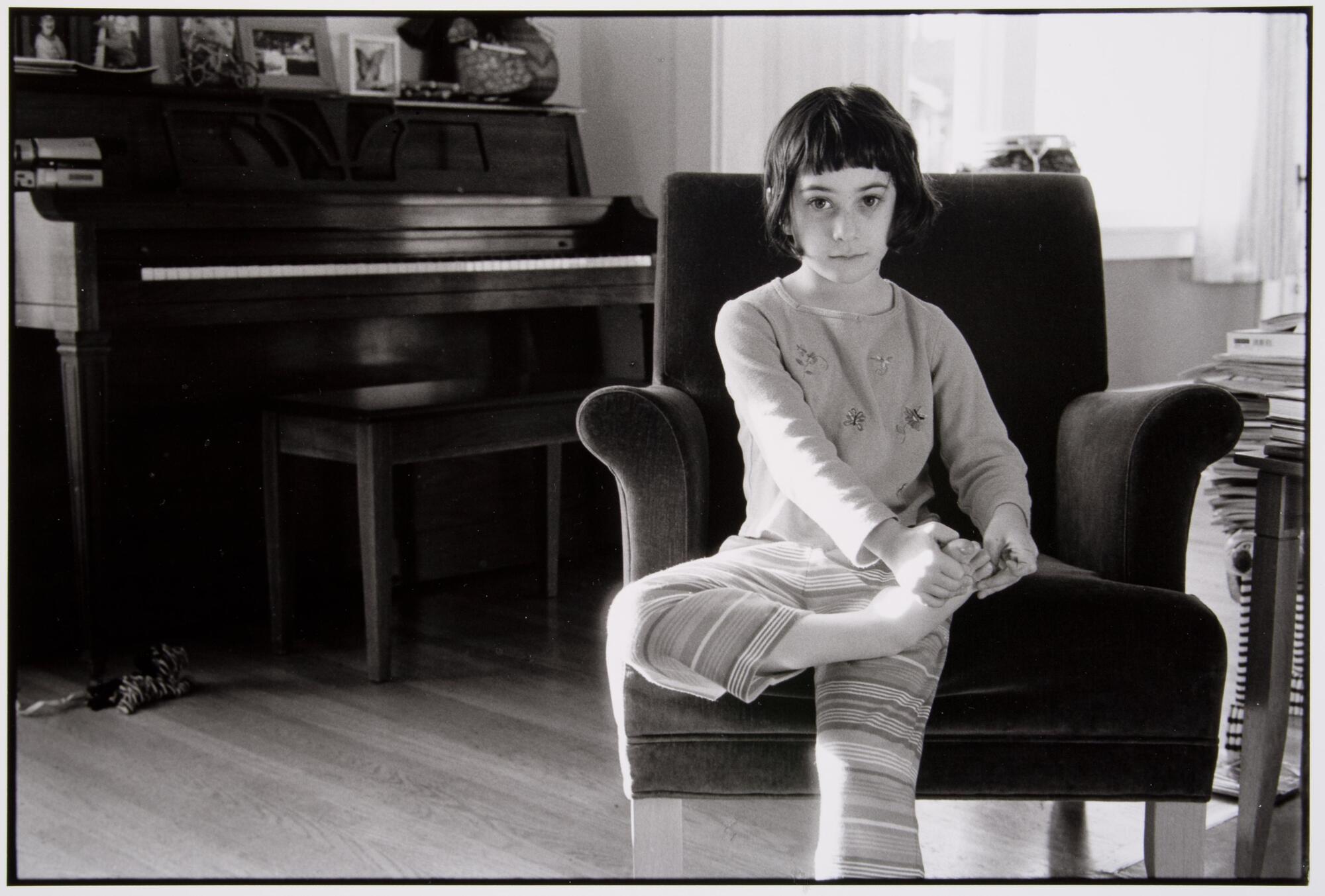 A girl with dark hair and heavy bangs is seated on a chair. Her left foot is brought up and being held by her hands. There is a piano in the background and wood floors.