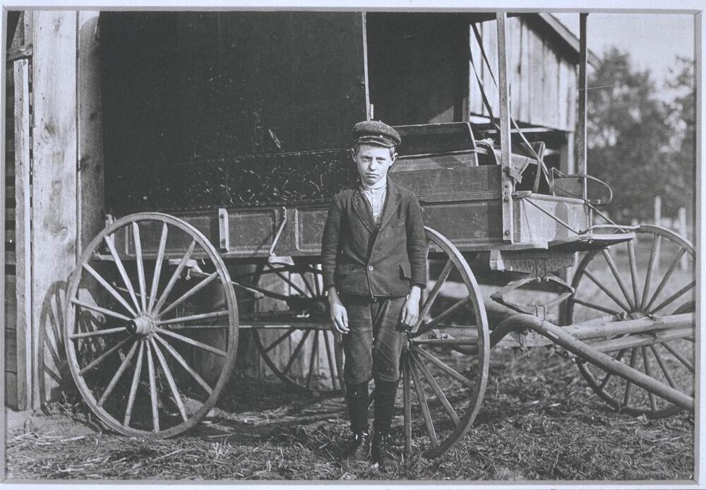This photograph depicts a view of a young boy standing by a covered wooden wagon. The back of the wagon rests against the side of a wooden building, its front is unhitched and resting on a ground strewn with hay.  