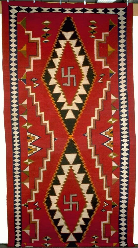 Red rug with black, white, yellow, and blue geometric designs. Two vertically oriented diamond shapes in the center. White and yellow stepped patterns moving towards the sides and blue and white triangular shapes forming a pattern as a border.