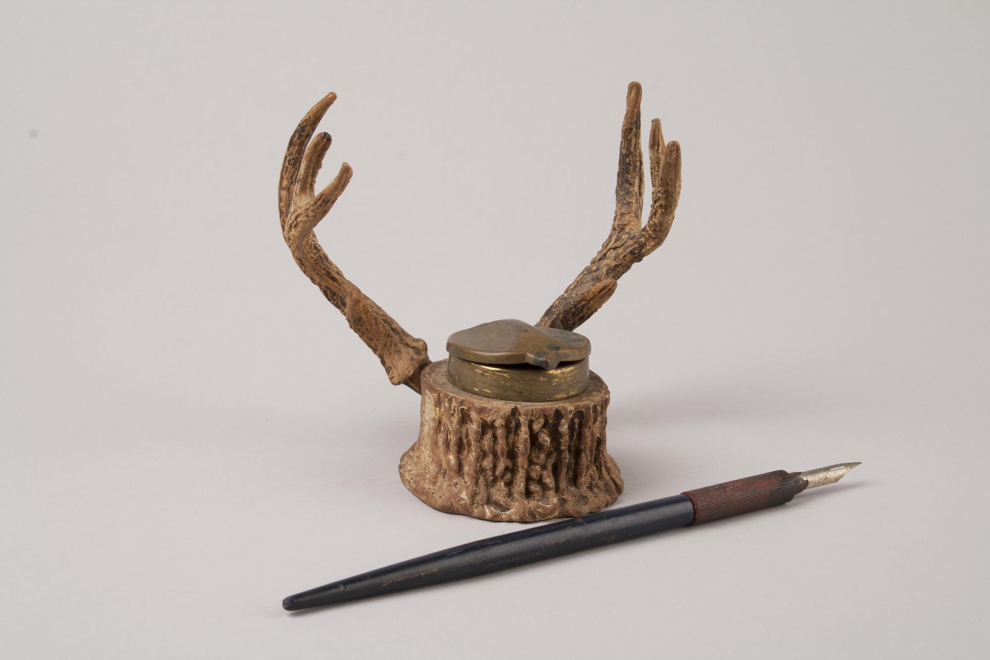 A small inkwell in a shape of a deer head has two deer horns of the left and the right. The two deer horns serve as an alter pen holder for the inkwell.