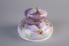 A purple color inkwell made with porcelain has drawings of apple blossom all around the outside of the inkwell. And there is a lid cover on the top of the inkwell.