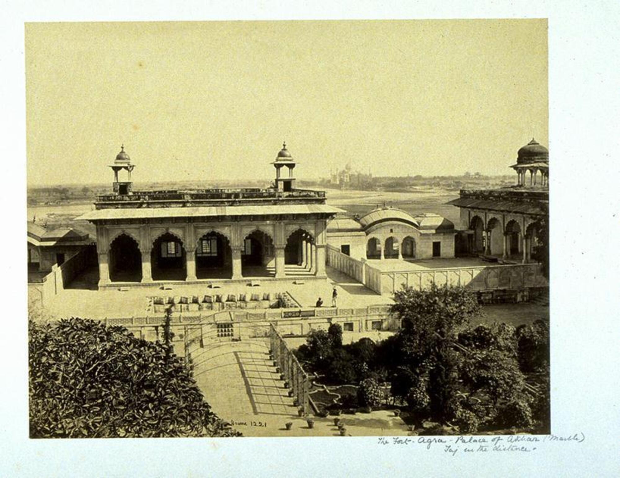 This photograph depicts a panorama view of a palatial structure with archways and two domed watchtowers on its roof. Before the structure stand two figures talking and a garden full of trees and potted plants.