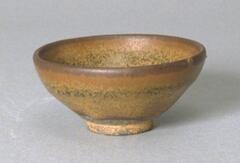 A conical bowl on a tall straight foot ring, covered in a dark brown-black glaze with intense and copious russet and ochre hare's fur markings (兔毫盏 <em>tuhao zhan</em>).