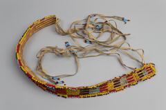 Beaded necklace made up of four brown cord wrapped coils with beaded detail in yellow, black and dark blue, light blue and red. Tie closure with four long leather tassels at each end. Tassles have beaded end details in mostly light blue with dark blue, red and yellow, white tag attached.