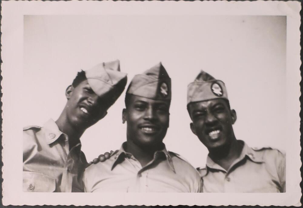 Three smiling men in uniform, photographed from the shoulders up, look slightly downward at the camera. The man on the right has his hand on the shoulder of the man in the middle.