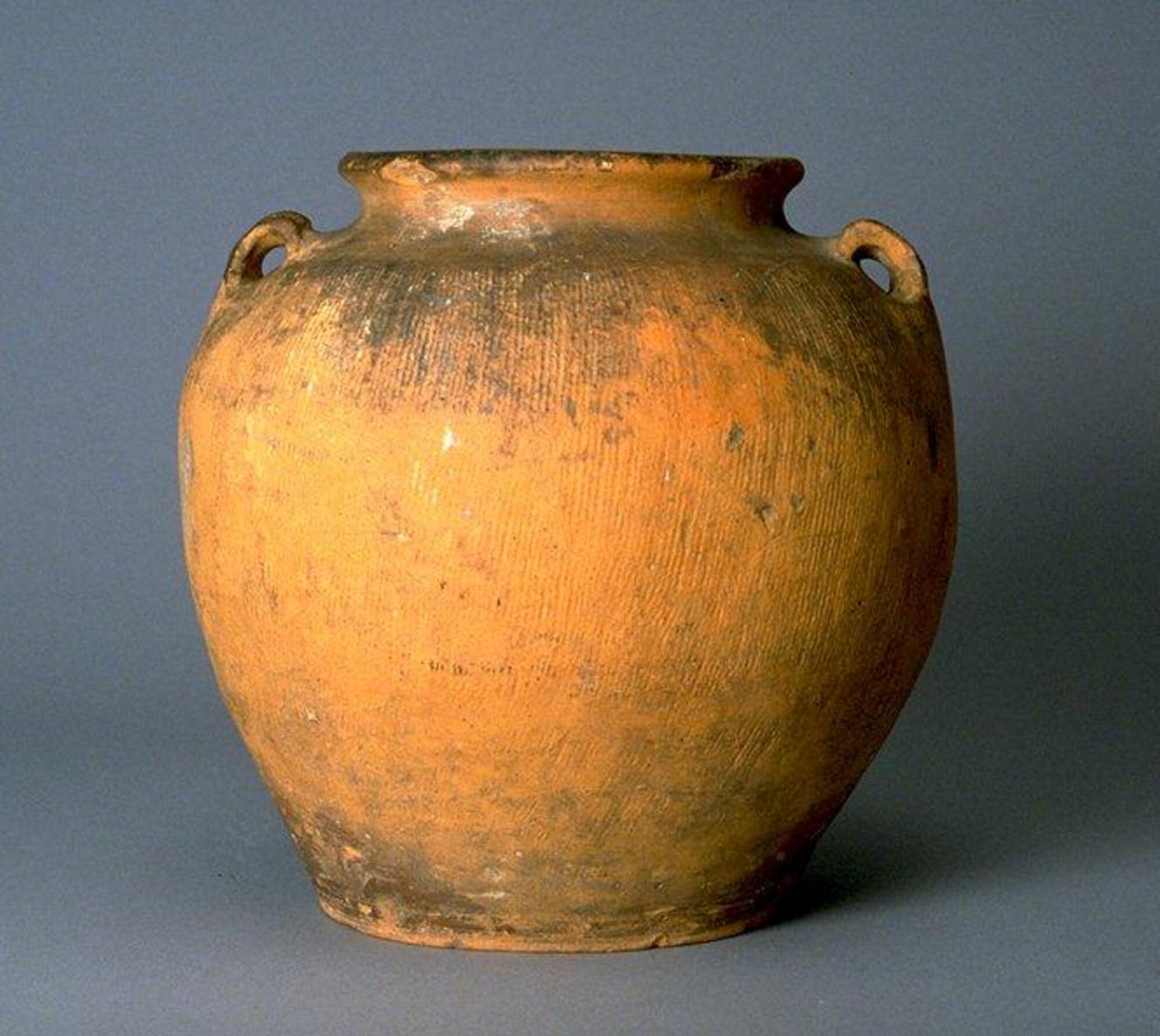 This unglazed, earthenware globular jar has a flat base, wide neck, and two lug handles applied to shoulder with a combed pattern around body.