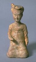 An earthenware figure of a kneeling woman wearing a long dress with an empire waist, her hair coiffed high upon her head, and arms raised as if once holding something.  It is covered in white slip and polychrome mineral pigment. 