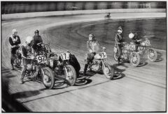 A photograph of six men straddling numbered motorcycles lined up at a starting line on a dirt race track. They wear motorcycle gear and interact with one another.