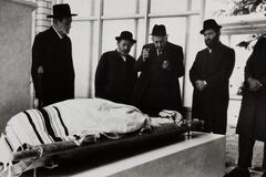 A group of Hassidic men standing over a body covered in a cloth. There are windows behind them.