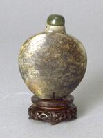 A brown, gold, green colored chicken bone jade and nephrite snuff bottle. It is rounded in shape and slightly thins towards the bottom which is flat. Incised on the surface of the snuff bottle are two peaches with leaves and vines surrounding them. It also has a green stopper and it sitting on a decorated wooden stand.