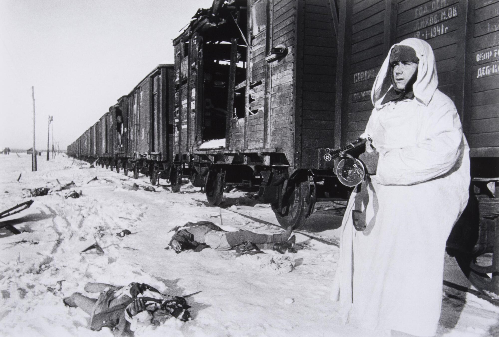 A soldier in winter gear holds a firearm next to an unmoving train while dead soldiers lie in the snow around him.